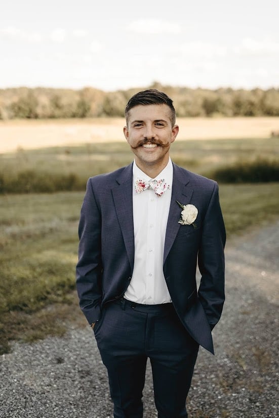 Man with distinctive mustache in formal navy suit with white boutonniere standing on rural road with fields behind